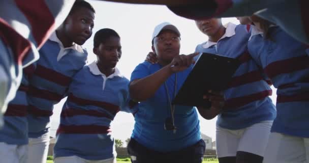 一群年轻的成年多种族女子橄榄球运动员站在橄榄球场上 手挽手拥抱着 一边听着她们的中年男女混合赛女教练的表演 一边准备一场橄榄球赛 一边背对着灯光 — 图库视频影像