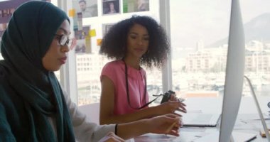 Modern bir ofiste çalışan iki genç melez iş kadınının yan görünüşü, bir kadın gözlük takıyor, tesettür takıyor ve bilgisayar monitörünün önünde oturuyor, diğer kadın ise bir kamera tutuyor.