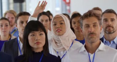 İş konferansında tesettürlü genç bir Asyalı iş kadınının ön planda el ele tutuşması ve soru sormak için ayağa kalkması, etrafındaki seyirciler arasında çeşitli iş adamlarının oturduğu görülüyor.