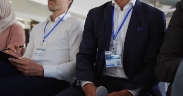 Nahaufnahme eines jungen kaukasischen Geschäftsmannes im Publikum einer Business-Konferenz, der die Hand hebt, aufsteht und ein Mikrofon benutzt, um eine Frage zu stellen, eine vielfältige Gruppe anderer Geschäftsleute
