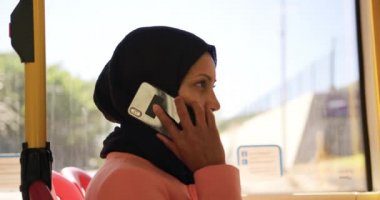 Bir şehirde bir otobüste oturan bir hijab commuting giyen genç bir karışık ırk kadın yan görünümü, akıllı telefon konuşurken