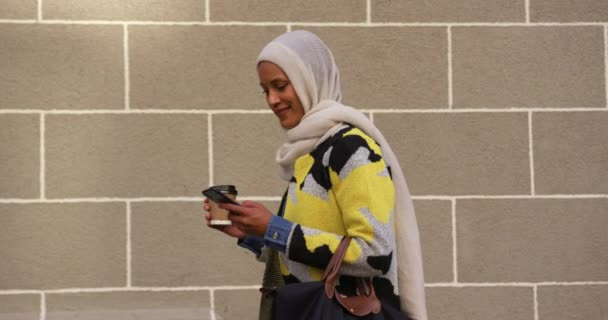 一个年轻的混血女人戴着头巾在城市街道上行走的侧视图 使用智能手机拿着咖啡杯 微笑着对着相机 — 图库视频影像