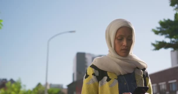 一个年轻的混血妇女戴着头巾在城市街道上行走 使用智能手机的前视图 — 图库视频影像