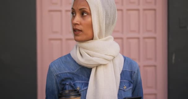 街の通りに立つヒジャーブを着た若い混合レース女性の正面図は コーヒーカップを持ち カメラに微笑むスマートフォンを使用して — ストック動画