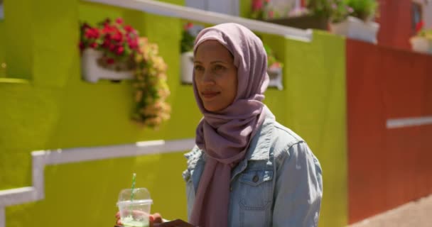 街の通りを歩くヒジャーブを着た若い混合人種女性の側面図 カラフルな家々を背景にジュースを飲む — ストック動画