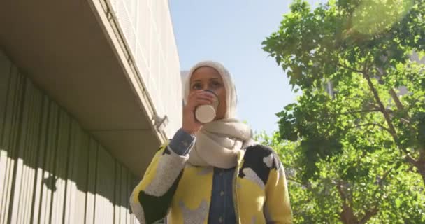 一个年轻的混血妇女穿着头巾走在城市街道 喝咖啡的前视图 — 图库视频影像