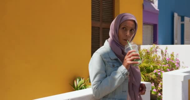 街の通りでヒジャーブを着てジュースを飲み カラフルな家を背景に壁に寄りかかっている若い混合人種女性の正面図 — ストック動画