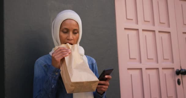 サンドイッチを食べるスマートフォンを使って 街の通りに立つヒジャーブを着た若い混合レース女性の正面図 — ストック動画
