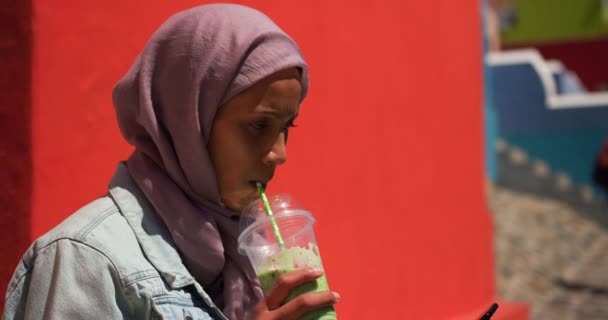 在城市街道上 一位穿着头巾的年轻混血妇女戴着头巾 喝着果汁 使用背景有红墙的智能手机的侧视图特写 — 图库视频影像