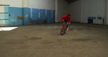 Terk edilmiş bir depoda hileler yaparken bmx bisikletinin arka tekerleğinde zıplayan, dönen ve dengelenen beyaz bir adamın yan görünümü