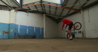 Terk edilmiş bir depoda hileler yaparken bmx bisikletinin ön tekerleğinde zıplayan, dönen ve dengelenen beyaz bir adamın yan görünümü