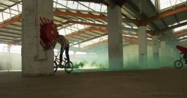 Terk edilmiş bir depoda Bmx bisikletlerine atlayan ve binen iki beyaz gencin yan görüntüsü.