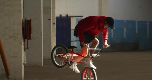 放棄された倉庫でトリックを練習しながら 日光のシャフトでBmxバイクの前輪にバランスをとる若い白人男性の側面図 — ストック動画