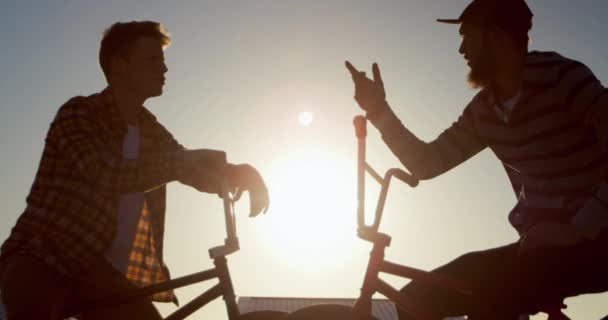放棄された倉庫の屋上で話しているBmxバイクに座っている2人の若い白人男性のサイドビュークローズアップ 夕日に照らされたバックライト — ストック動画
