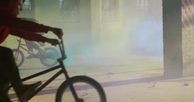Terk edilmiş bir depoda, mavi ve sarı sis bombaları ile Bmx bisiklet sürme ve hile yapan iki genç Kafkas erkeklerin yan görünümü