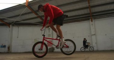Terk edilmiş bir depoda hileler yaparken bmx bisikletinin ön tekerleğinde zıplayan, dönen ve dengelenen beyaz bir adamın yan görünümü