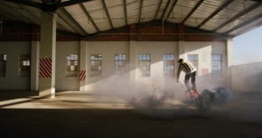 Terk edilmiş bir depoda, üzerinde gri bir sis bombası olan Bir Bmx bisikletinin üzerine atlayan beyaz bir adamın yan görüntüsü