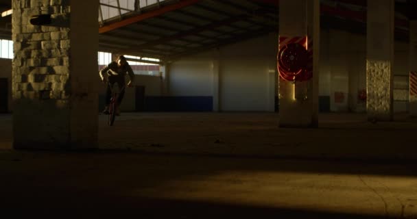 Bmxバイクに乗って 放棄された倉庫でトリックをやっている若い白人男性の側面図 — ストック動画