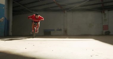 Terk edilmiş bir depoda, güneş ışığının altında Bmx motoruna atlayan genç bir beyaz adamın ön görüntüsü.