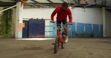 Terk edilmiş bir depoda numaralar çalışırken Bmx motorunun ön tekerleğinde atlayan, dönen ve dengede duran genç bir beyaz adamın ön görüntüsü.