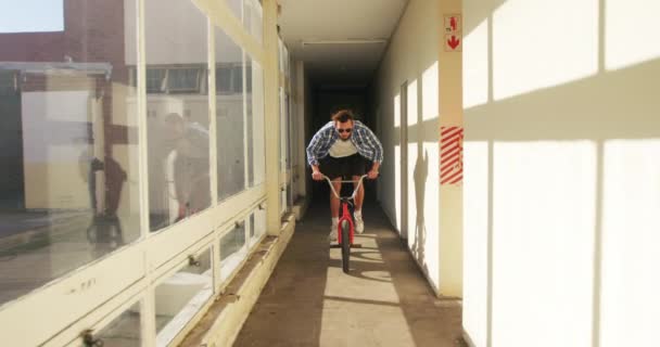 太陽の下で放棄された倉庫の狭い廊下でBmxバイクに飛び乗るサングラスをかけた若い白人男性の正面図 — ストック動画