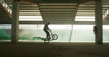 Terk edilmiş bir depoda, üzerinde yeşil bir duman bombası olan Bmx motosikletine atlayan genç bir beyaz adamın yan görüntüsü, ön planda başka bir sihouetted Bmx sürücüsü geçiyor.