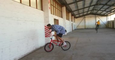 Bmx bisikleti süren ve terk edilmiş bir depoda duvarda koşan beyaz bir adamın arka planda yaptığı numaralar ön planda.