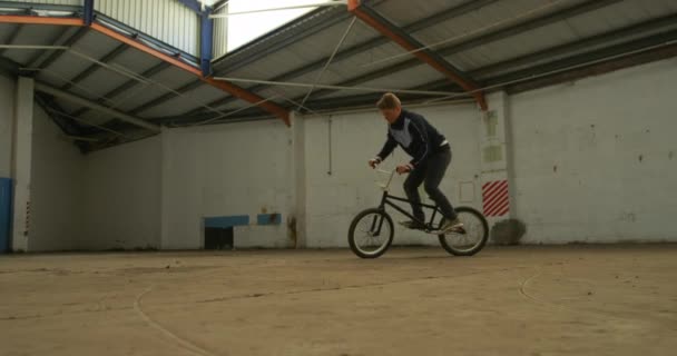 放棄された倉庫でトリックを練習しながら 若い白人男性がジャンプし Bmx自転車をオンにするの側面図 — ストック動画