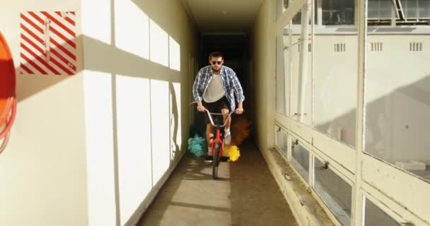 一个戴着太阳镜的白人年轻人骑着一辆带着绿色和橙色烟雾弹的Bmx自行车 在一个废弃的仓库里跳到一个狭窄的走廊里晒太阳 — 图库视频影像