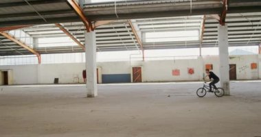 Terk edilmiş bir depoda Bmx bisikleti süren genç bir beyaz adamın yan görüntüsü.