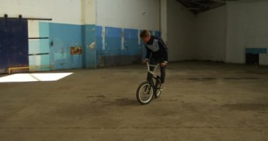 Terk edilmiş bir depoda, Bmx bisikleti üzerinde hoplayıp zıplayan beyaz bir adamın yan görüntüsü.