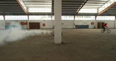 Terk edilmiş bir depoda, üzerinde gri bir duman bombası olan Bmx bisikletiyle atlayan genç bir beyaz adamın yan görüntüsü.