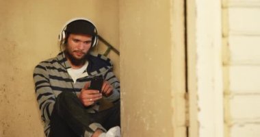 Beyzbol şapkası ve kulaklık takmış genç beyaz bir adamın, terk edilmiş bir depoda duvara yaslanmış, elinde akıllı bir telefonla müzik dinlediği ön görüntüsü.