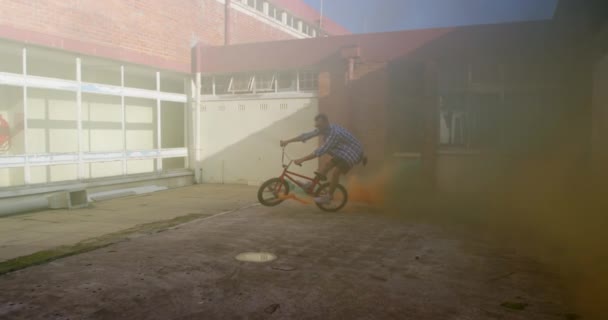 青とオレンジの煙の手榴弾がそれに接続されているBmx自転車に飛び乗る若い白人男性の側面図放棄された倉庫の外 — ストック動画