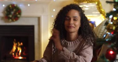 Noel 'de oturma odasında oturan mutlu, melez bir kadının portresi, kameraya gülümseyen ve gülen, Noel süsleri ve arka planda şöminede yanan alevlerle birlikte.