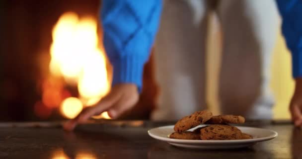 Vue de face de la section médiane d'une jeune fille métissée assise dans son salon à Noël et prenant un biscuit sur la table dans son salon, avec les flammes scintillantes d'un feu ouvert dans la cheminée en arrière-plan 