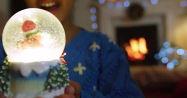 Noel 'de oturma odasında oturmuş, gülümseyen, kucaklayan ve parlayan kar küresine bakan melez genç bir kızın ön görüntüsü.