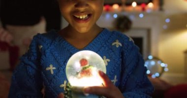 Noel 'de oturma odasında oturmuş, gülümseyen, kucaklayan ve parlayan kar küresine bakan melez genç bir kızın ön görüntüsü.
