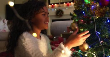 Noel 'de oturma odasında oturan, Noel ağacını süsleyen ve sevinçle gülümseyen melez bir kızın yan görüntüsü.