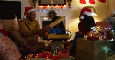 Noel 'de küçük kızlarıyla birlikte oturma odalarında, yerde yan yana oturan, Noel Baba şapkası takan bir çift ve onlardan hediye açan bir kız ile melez bir ailenin ön görüntüsü.