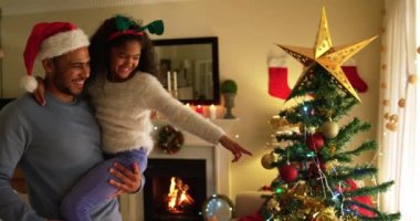 Noel 'de küçük kızlarıyla oturma odalarında oturan melez bir ailenin yan görüntüsü. Adam Noel Baba şapkası takıyor ve kızını kucağında tutuyor.