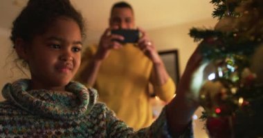 Düşük açılı bir bakış açısıyla melez bir baba ve genç kızı Noel 'de oturma odalarında otururken kızının Noel ağacını süslerken fotoğrafını çeken adam ve her ikisi de gülüyor.