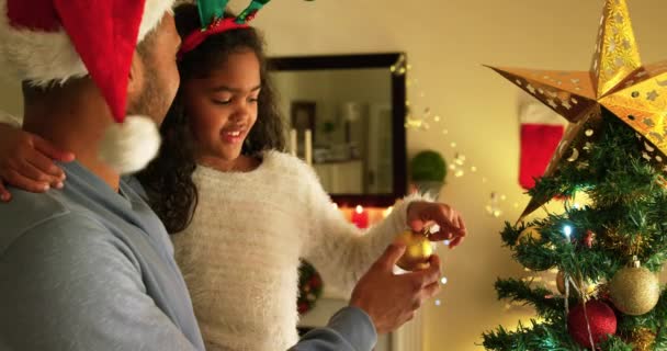 在圣诞节那天 一位混血的父亲和他年幼的女儿坐在客厅里 一边看一边看 这位男子戴着一顶圣诞老人帽 抱着他的女儿装饰着圣诞树 — 图库视频影像