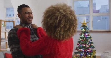 Arka planda dekore edilmiş bir Noel ağacı ile bir dairenin oturma odasında kucaklayan genç bir karışık ırk adam ve kadın yan görünümü