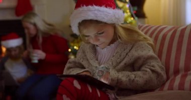 Yan görüntüde, Noel Baba şapkası takmış genç beyaz bir kız Noel zamanı oturma odasında tablet bilgisayar kullanıyor, annesi ve erkek kardeşi şöminenin arkasında oturuyor.