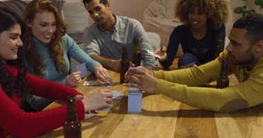 Masanın etrafında oturan, kağıt oynayan ve bira içen beş genç erkek ve kadın arkadaşından oluşan bir grubun ön görüntüsü.