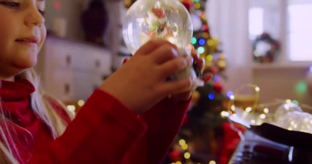 クリスマスの時間にシッティングルームで雪の地球を保持し 振る若い白人の女の子の近くにサイドビューとそれを見て 背景に装飾されたクリスマスツリー — ストック動画