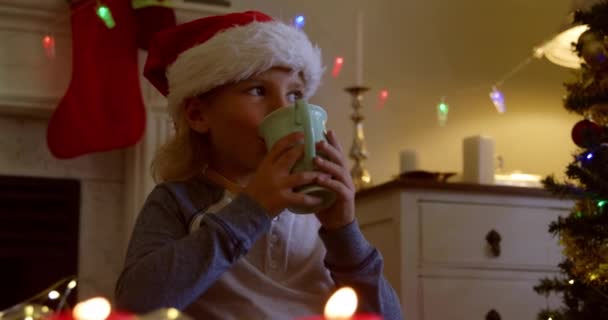 サンタの帽子をかぶった若い白人の少年がクリスマスの時に座っている部屋でカップを持って飲み 背景に装飾が施され 前景にろうそくが灯されています — ストック動画