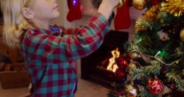 Genç beyaz bir çocuğun Noel ağacını Noel zamanı oturma odasında takılarla süslemesinin yan görüntüsü.