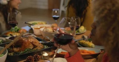 Bir grup yetişkin ve etnik çeşitlilikte genç kız arkadaşın Noel yemeği için evlerinde düşük bir masada oturup kadeh kaldırırken üçünün Noel Baba şapkası taktığını görüyorum.